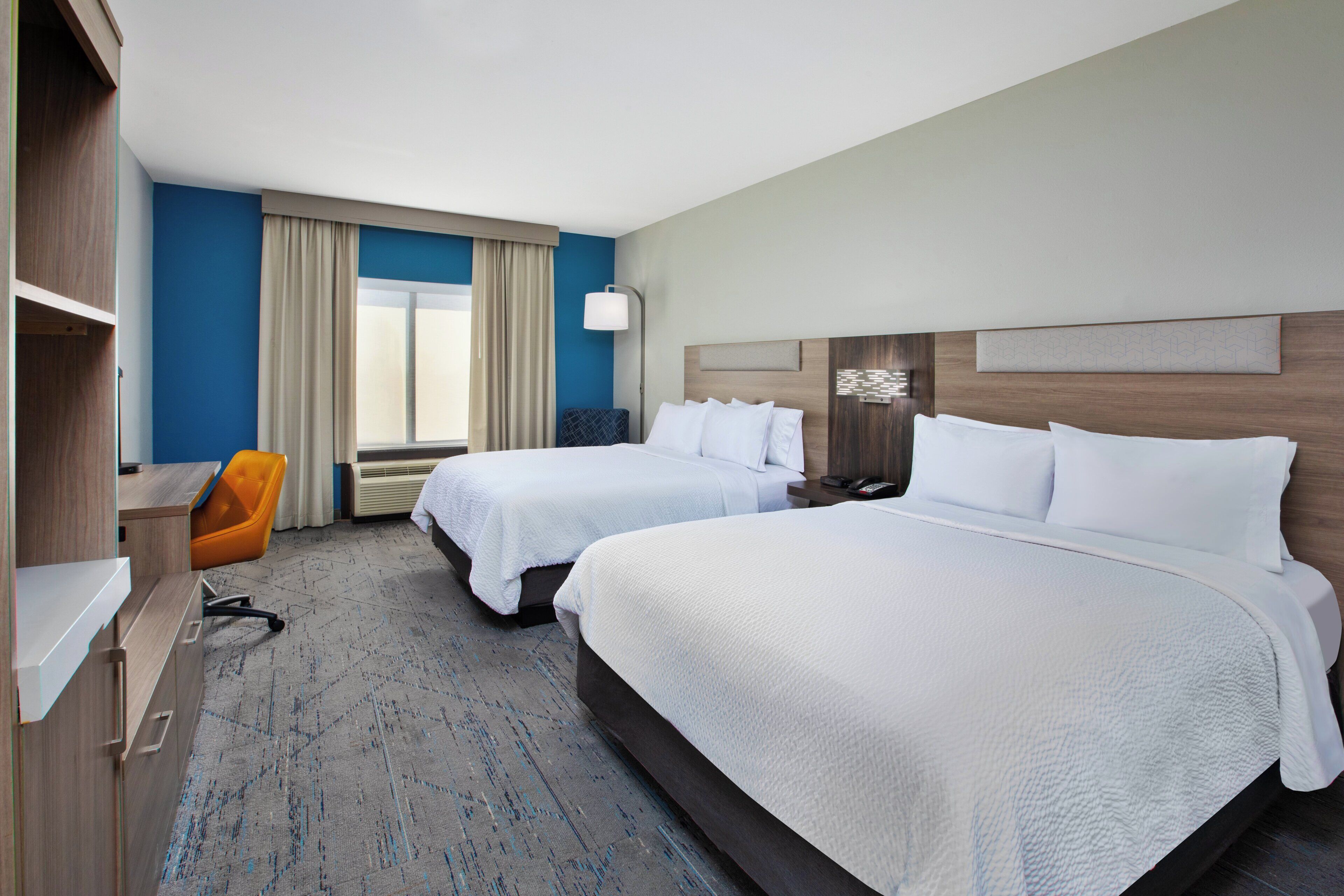 A hotel room with two beds, a desk, an orange chair, and a large window with curtains.