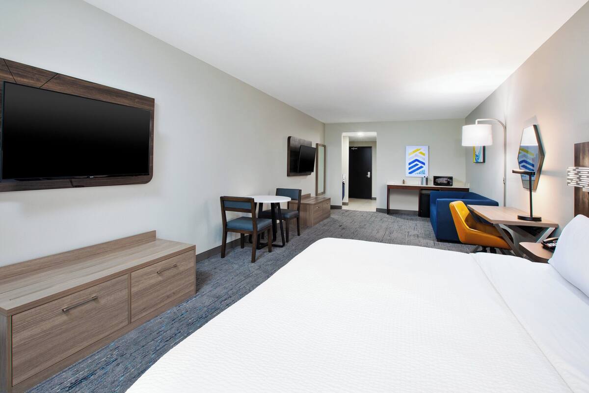 A hotel room with a large bed, a desk with a chair, a TV mounted on the wall, and a small seating area with a table and chairs.