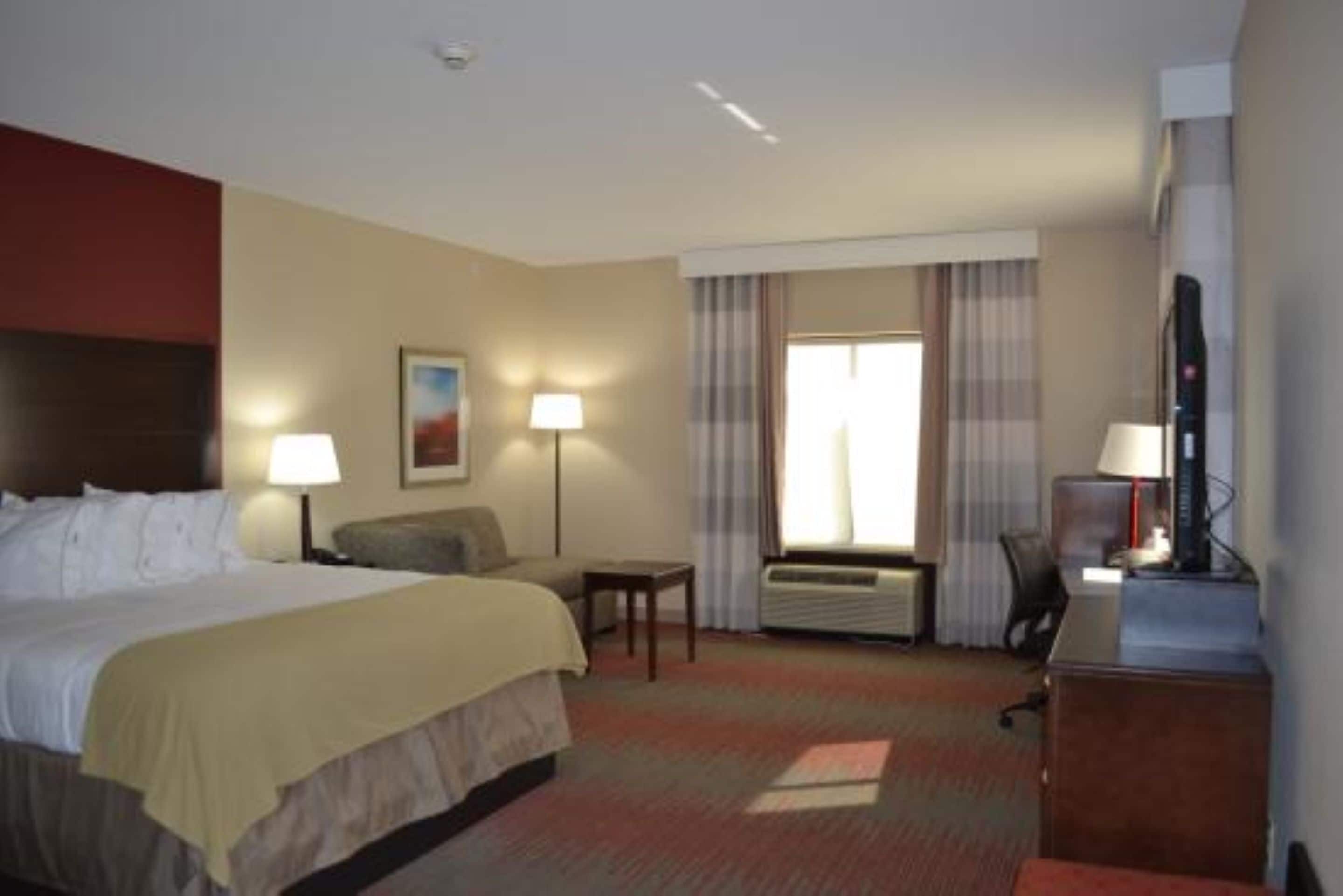 A hotel room with a bed, a desk, a chair, a lamp, a picture on the wall, and a window with curtains.