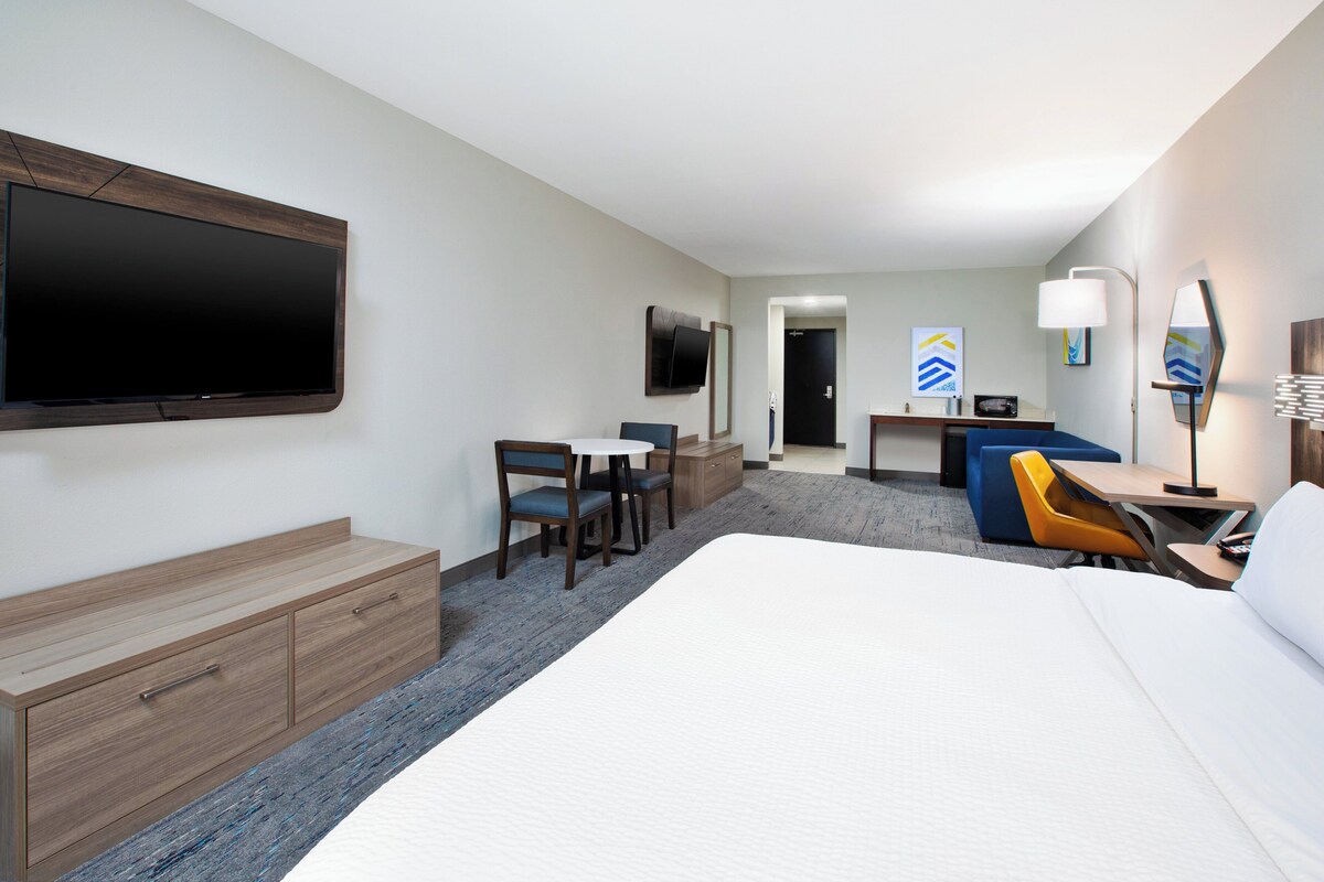A hotel room with a large bed, a desk with a chair, a TV mounted on the wall, and a small seating area with a table and chairs.