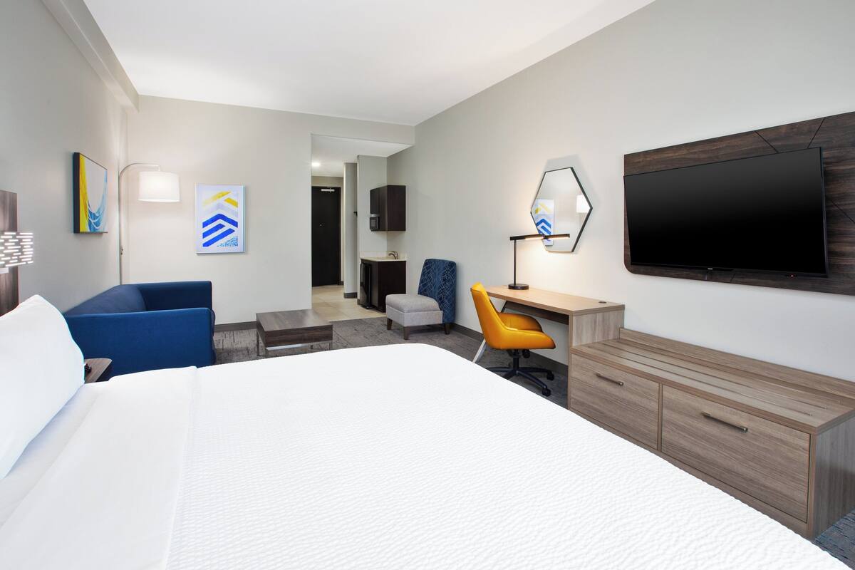 A hotel room with a large bed, a desk with a chair, a TV mounted on the wall, and artwork on the walls.
