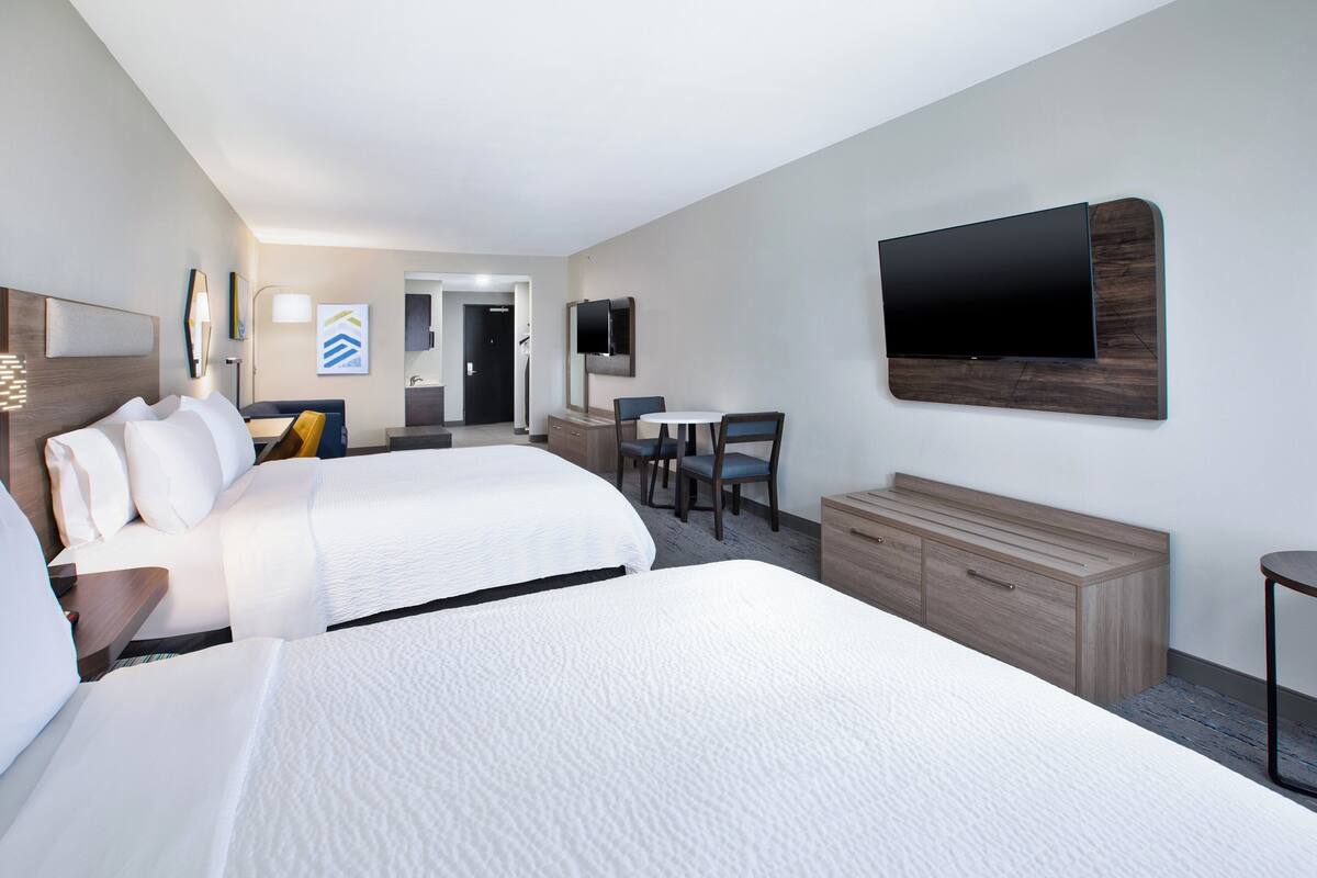 A hotel room with two beds, a desk, a chair, a TV mounted on the wall, and a bathroom visible through an open door.