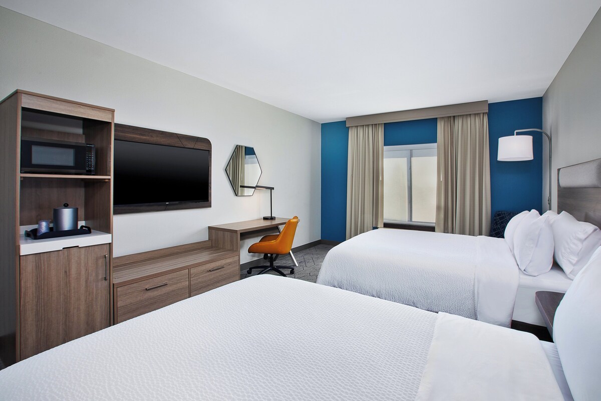 Habitación de hotel con dos camas, un escritorio con silla, televisor de pantalla plana, microondas y una ventana grande con cortinas.