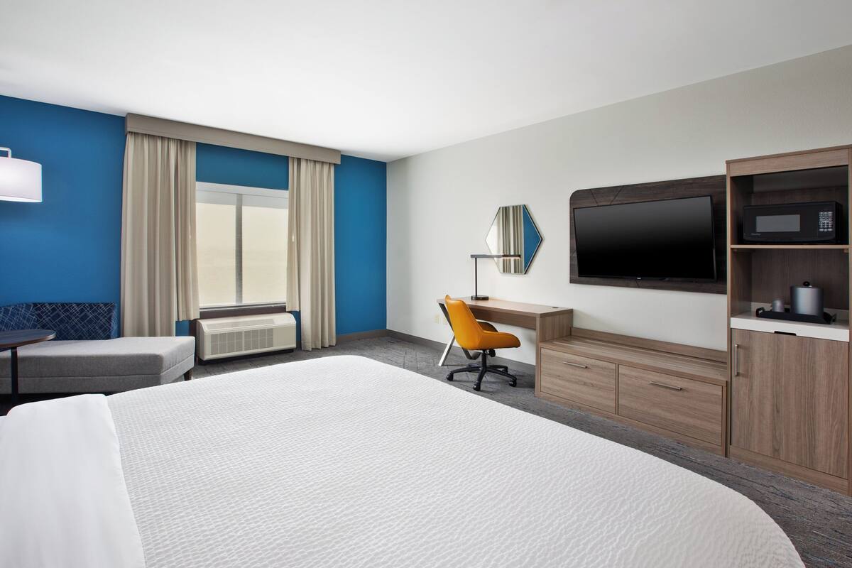 A hotel room with a large bed, a desk with a chair, a television, and a minibar.