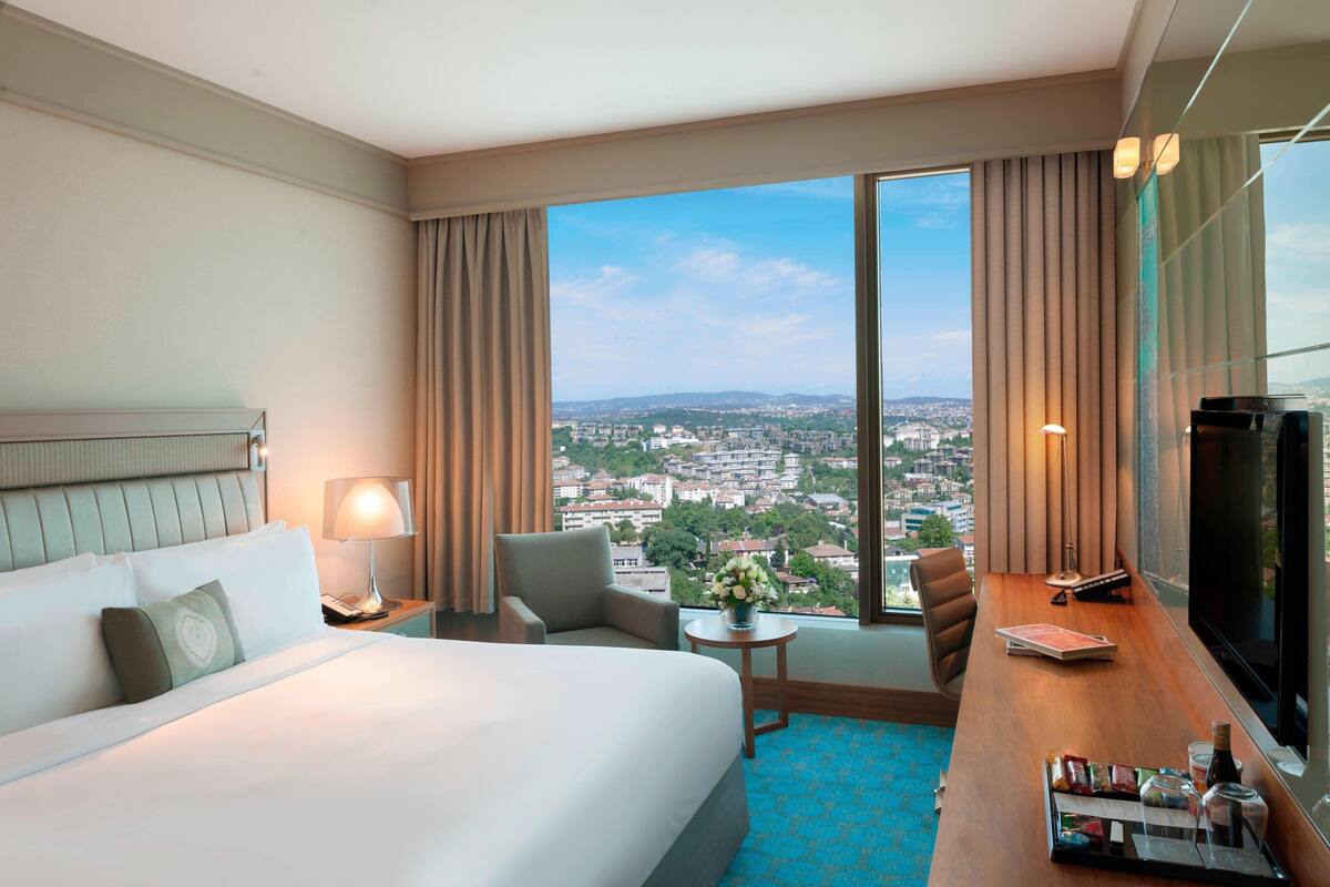 A hotel room with a large bed, a desk with a TV, a chair, a small table with a lamp, and a view of the city through large windows.