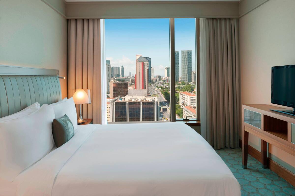 A hotel room with a large bed, a TV, a desk, and a city view through the window.