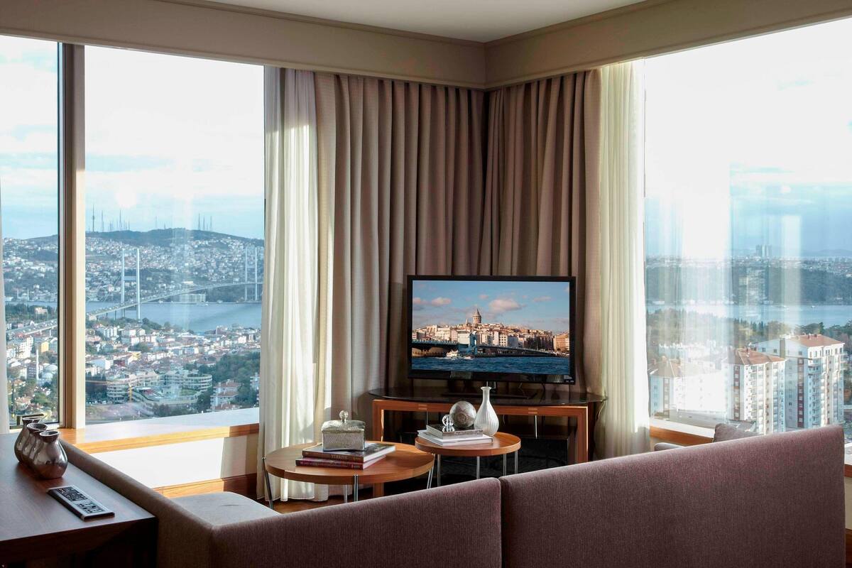 A hotel room with a large window offering a city view, a flat-screen TV, a sofa, a small table with books and a vase, and a remote control on the side table.