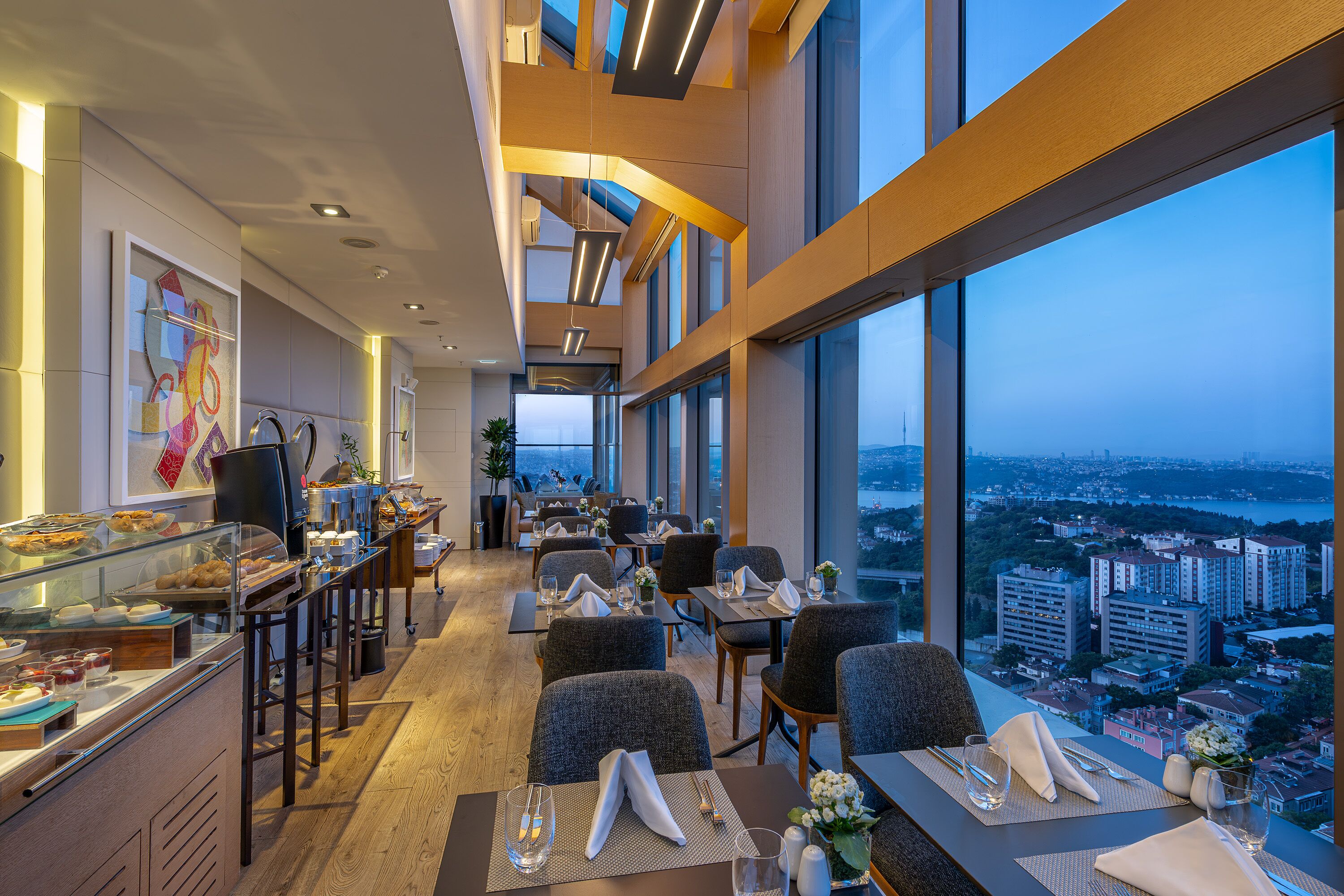 A modern hotel dining area with a city view, featuring a buffet, seating arrangements, and a large window overlooking the cityscape.