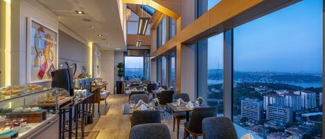 A modern hotel dining area with a city view, featuring a buffet, seating arrangements, and a large window overlooking the cityscape.