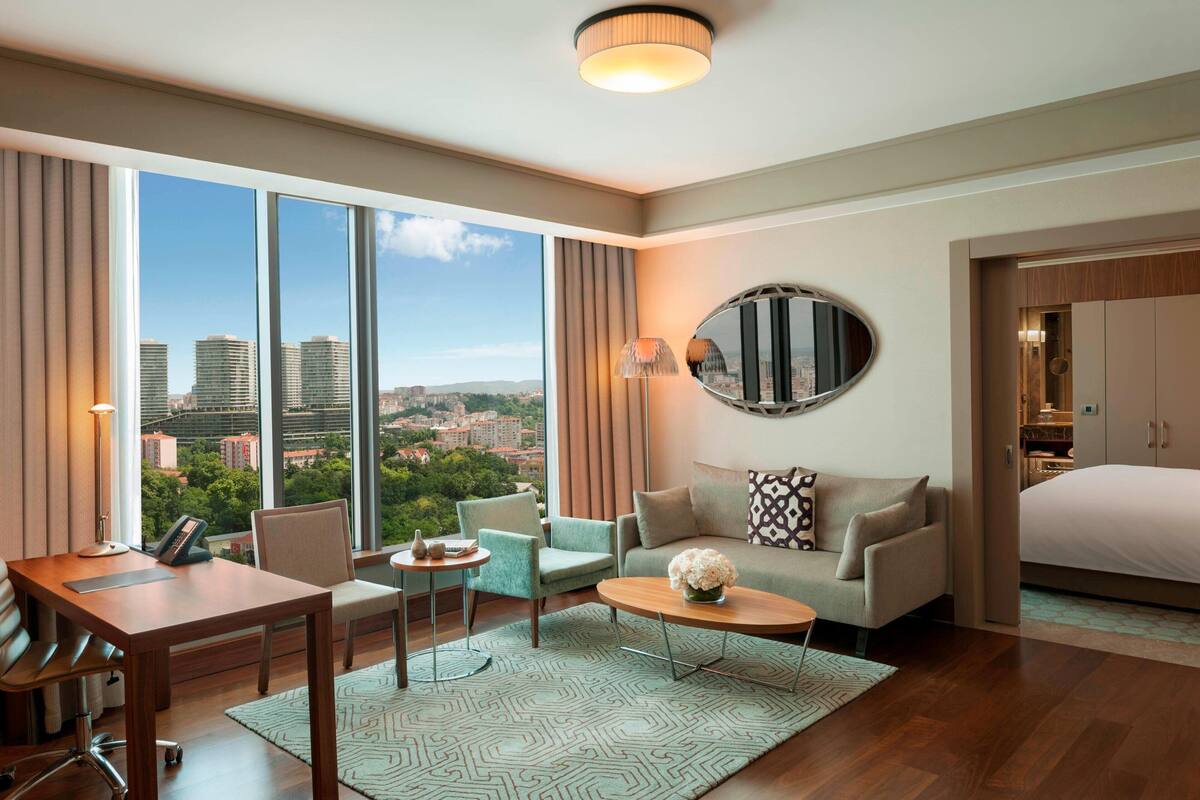 A modern hotel room with a large window offering a city view, a cozy seating area with a sofa and armchairs, a wooden desk with a chair, and a bed with a wooden headboard.