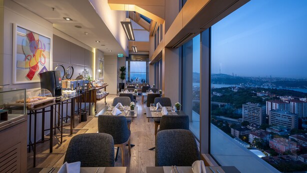 A modern restaurant with a city view, featuring a long counter with a coffee machine, seating area with tables and chairs, and a large window overlooking the cityscape. - Renaissance Istanbul Polat Bosphorus Hotel (Istanbul)