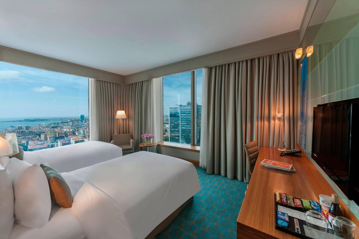 A hotel room with two beds, a desk with a TV, a window with curtains, and a view of the cityscape.