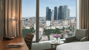 A hotel room with a large window offering a city view, a desk with a book, a chair, and a small table with a vase of flowers. - Renaissance Istanbul Polat Bosphorus Hotel (Istanbul)
