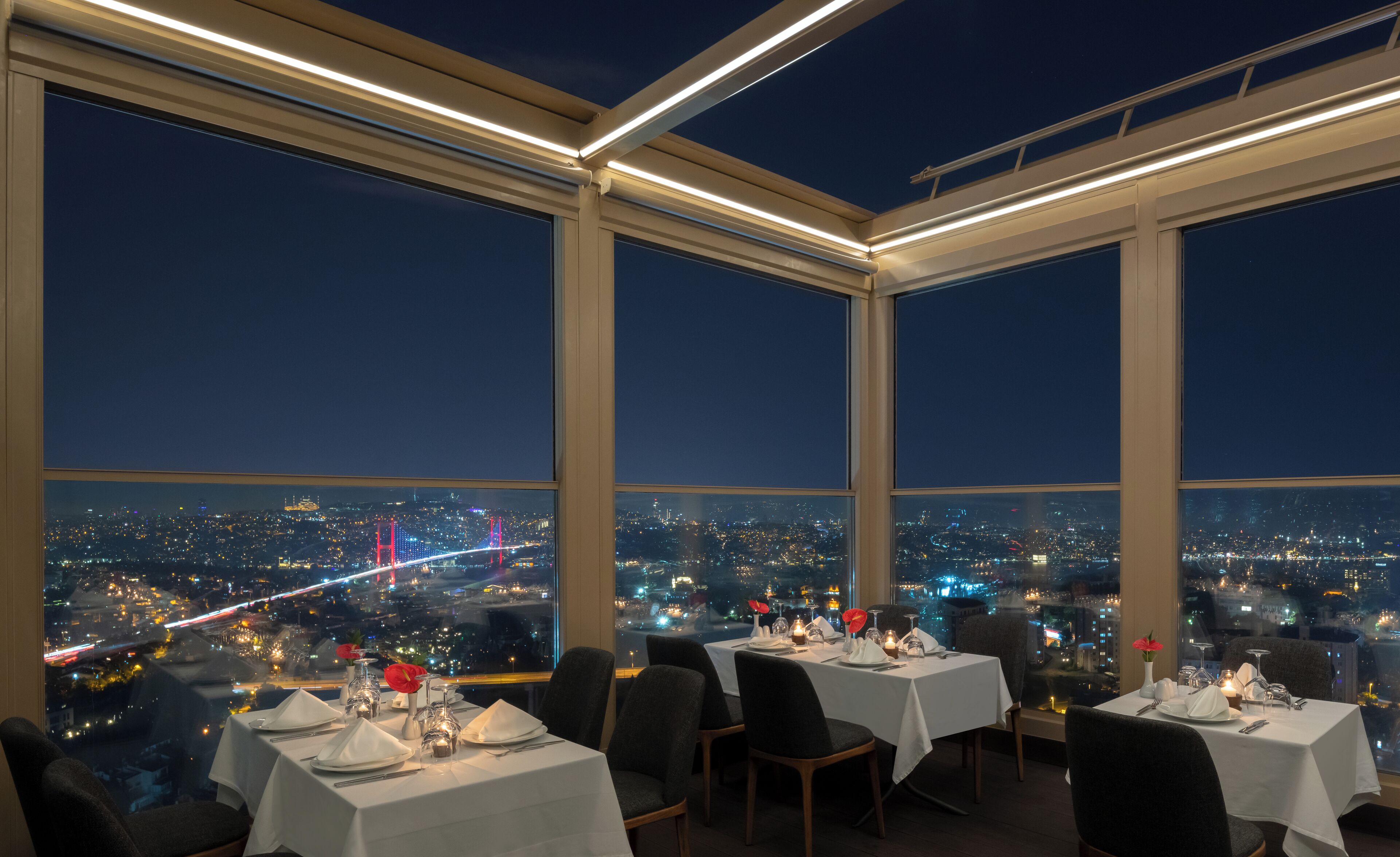 A dining area with tables set for two, large windows offering a cityscape view at night.