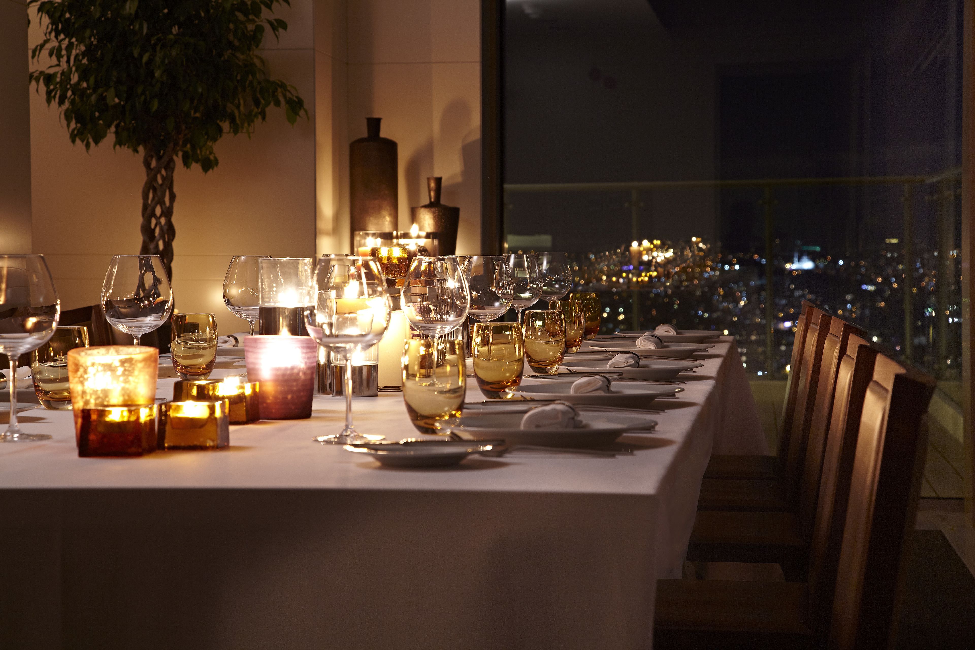 A dining table set for a meal with candles, wine glasses, and plates, overlooking a cityscape at night.