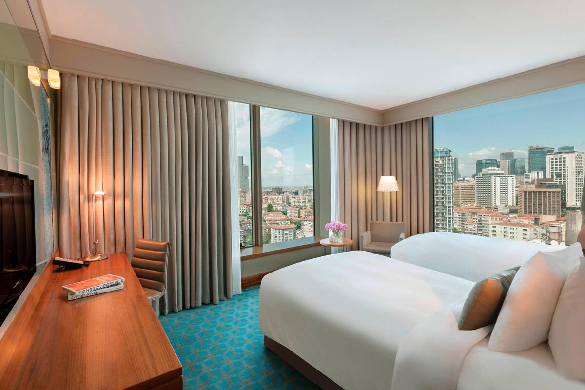 A hotel room with a large bed, a desk with a chair, a TV, and a view of a cityscape through large windows.