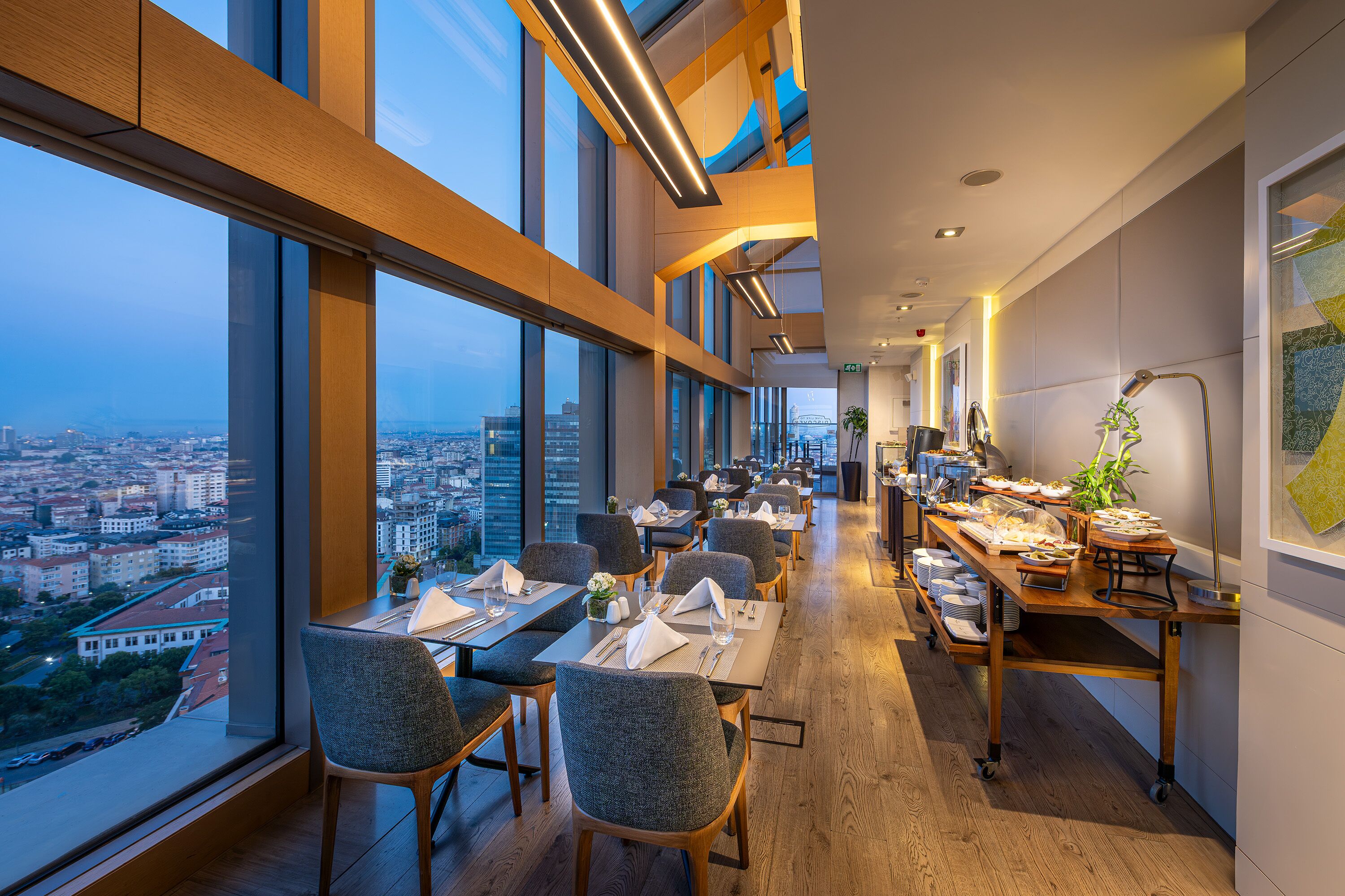 A modern dining area with high ceilings, large windows, and a city view.
