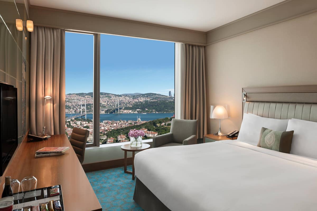 A hotel room with a large bed, a desk with a chair, a small table with a vase of flowers, and a view of a cityscape through large windows.