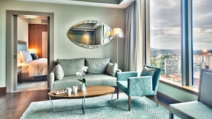 Quarto de hotel moderno com uma janela grande, um espelho redondo, um sofá, uma mesa de centro e vista para a paisagem urbana.