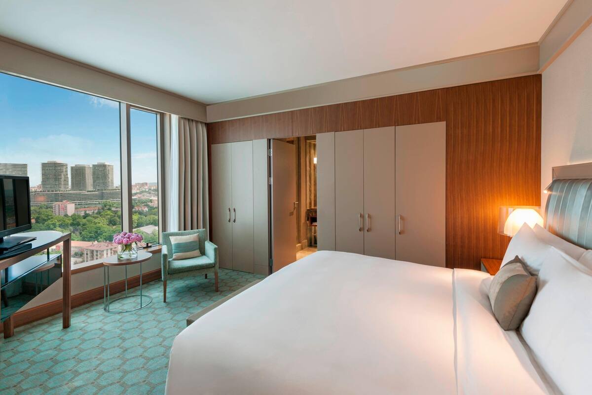 A hotel room with a large bed, a desk with a TV, a chair, and a view of the cityscape through large windows.