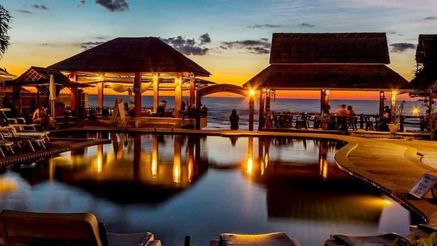 Uma área à beira da piscina com espreguiçadeiras e guarda-sóis, um espaço coberto para sentar e vista para o oceano ao pôr do sol.