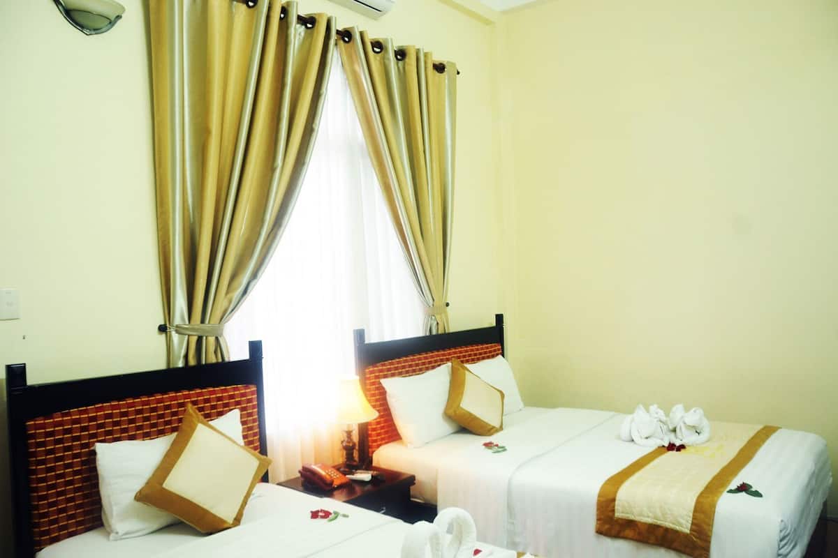 A hotel room with two beds, a window with curtains, a bedside table with a lamp, and a wall-mounted air conditioner.