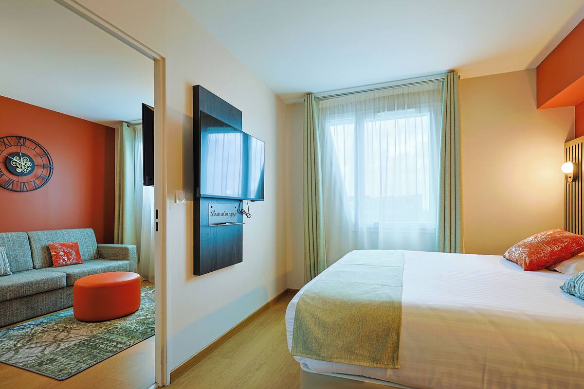 Een hotelkamer met een bed, een bank, een aan de muur bevestigde televisie en een raam met gordijnen.