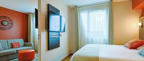 Una habitación de hotel con cama, sofá, televisor montado en la pared y ventana con cortinas.
