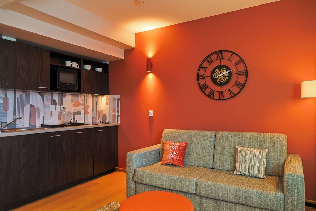 Een moderne woonkamer met een bank, een wandklok en een keuken met kasten en een magnetron.