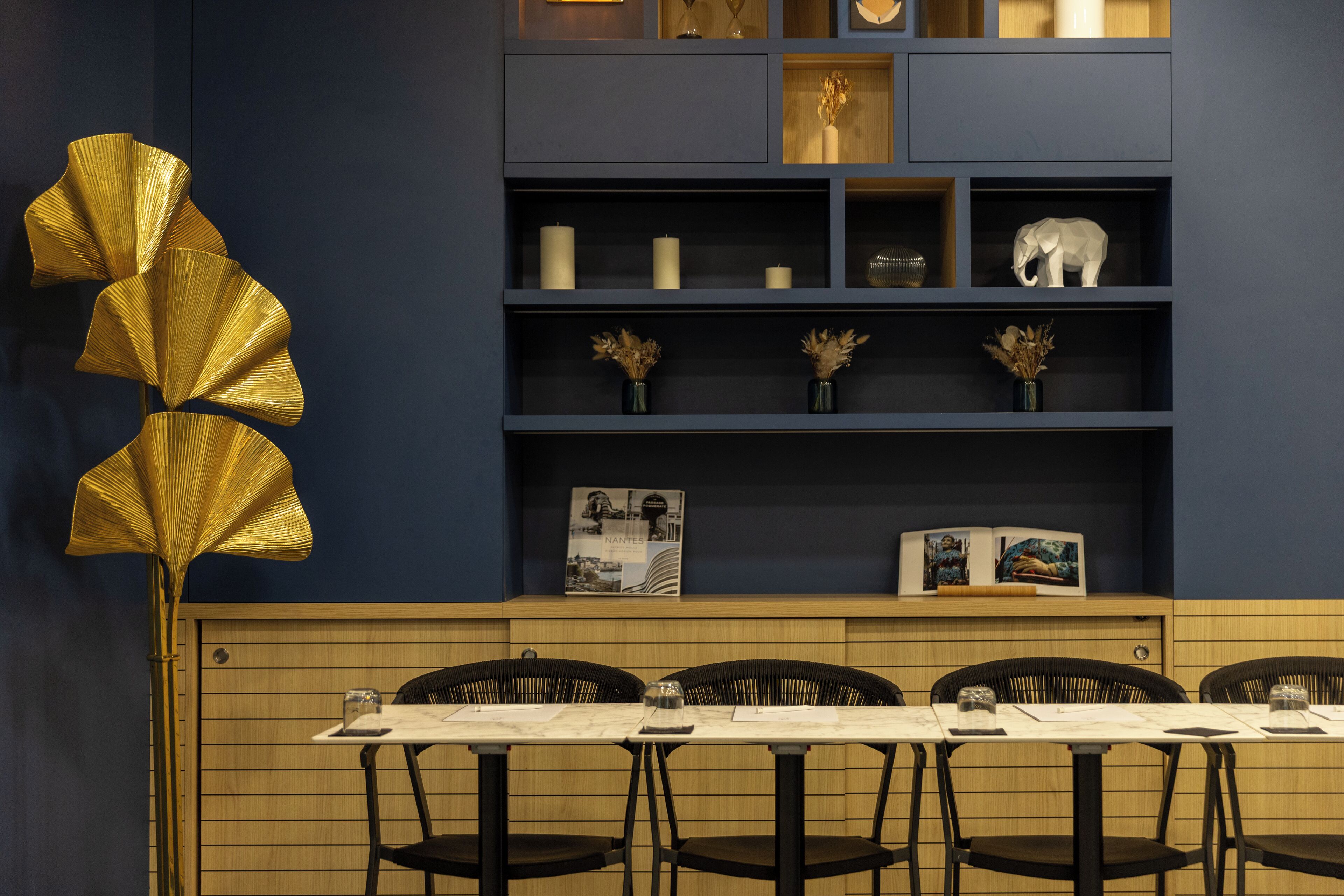 A dining area with a long table, black chairs, and a decorative golden fan. Shelves above the table hold candles, a vase with dried flowers, and a small elephant sculpture.