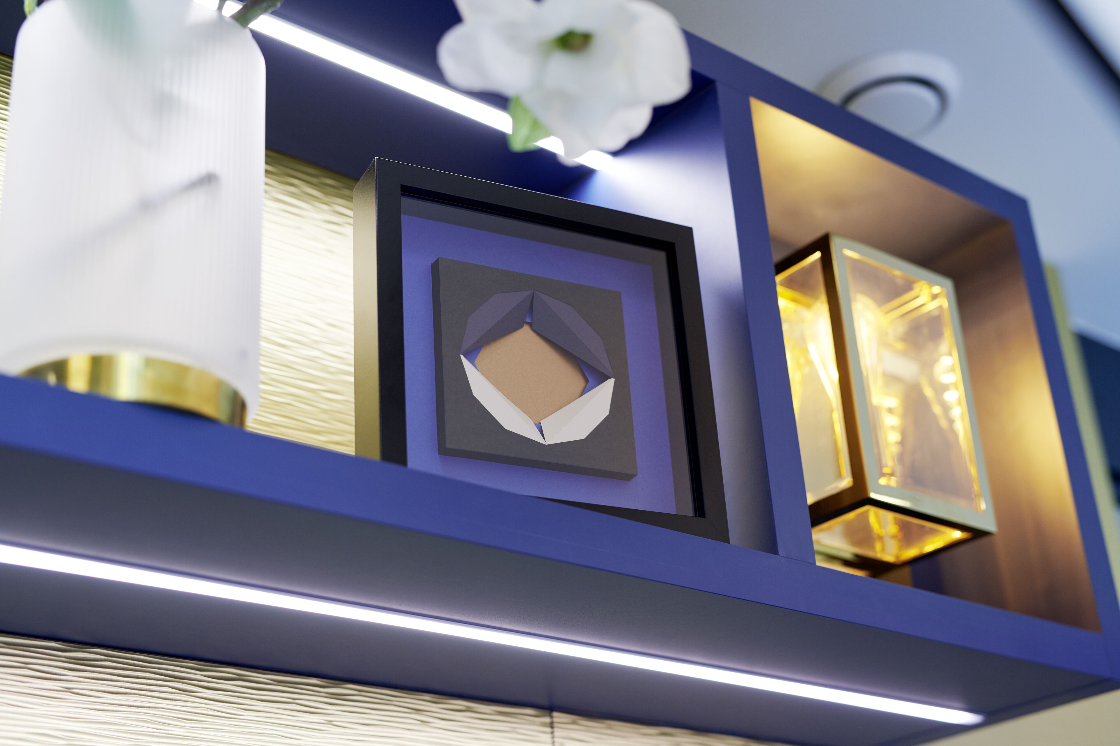 A framed artwork with a geometric design, a white orchid, and a lit wall-mounted lamp on a blue shelf.
