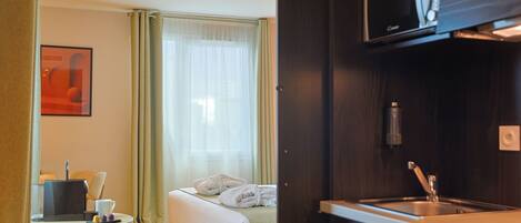 Une chambre d’hôtel comprenant un lit, un bureau avec un ordinateur portable, une chaise, une fenêtre avec des rideaux et une kitchenette équipée d’un four à micro-ondes et d’un évier.