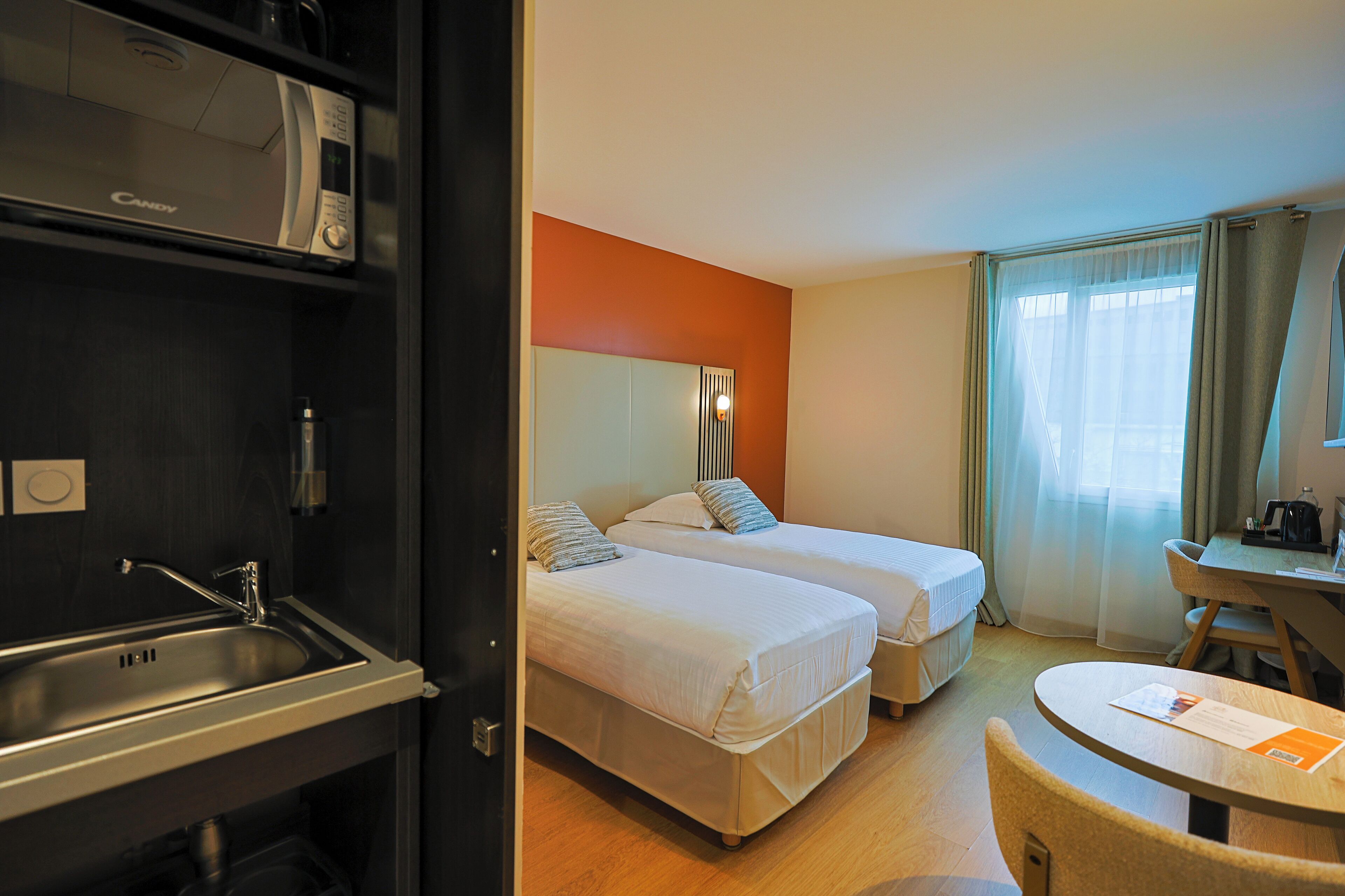 Une chambre d’hôtel avec deux lits, un bureau, une chaise, un lavabo et une fenêtre avec des rideaux.