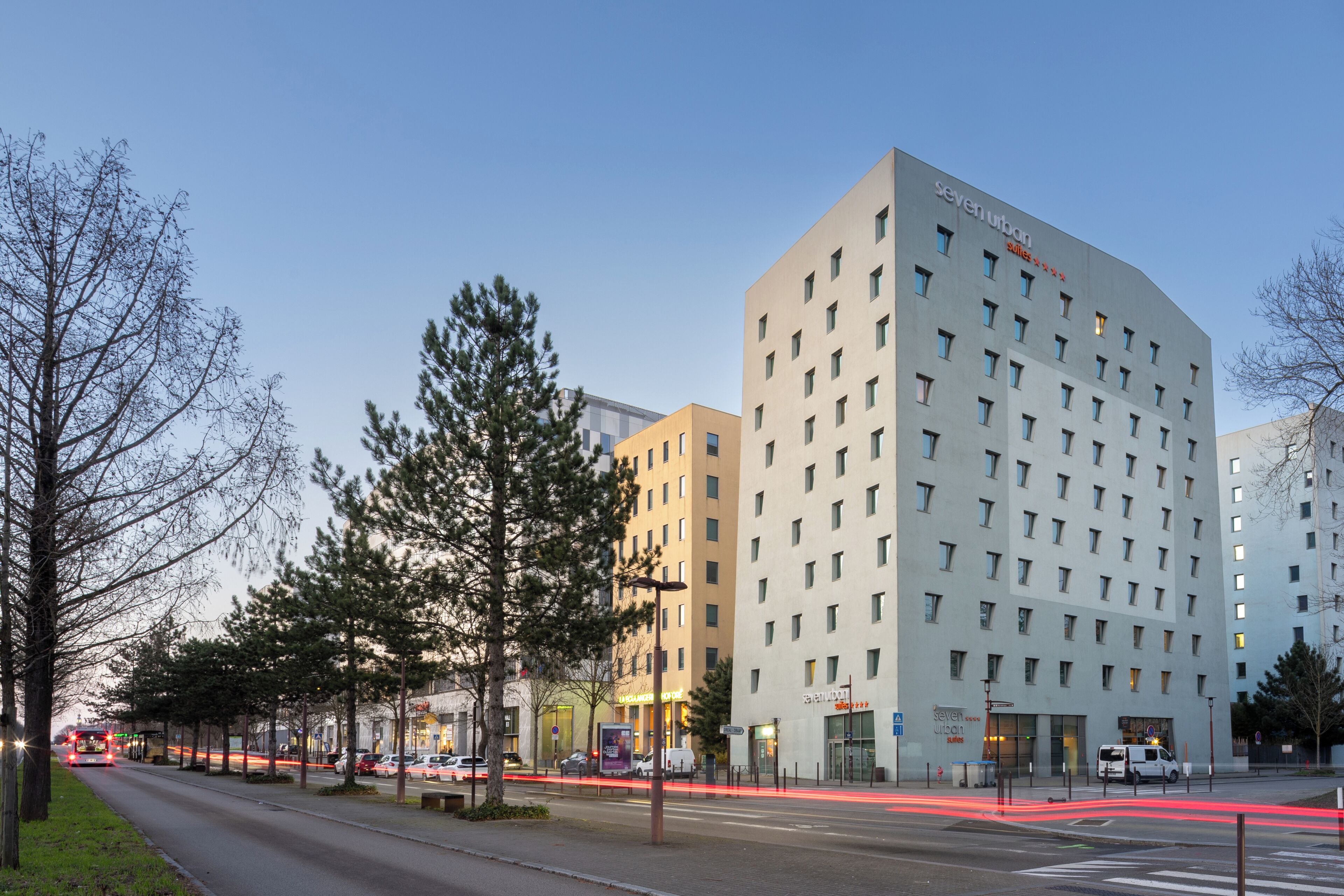 Un hôtel moderne à plusieurs étages, avec une enseigne bien visible indiquant « seven urban », et une vue dégagée sur la rue au premier plan.