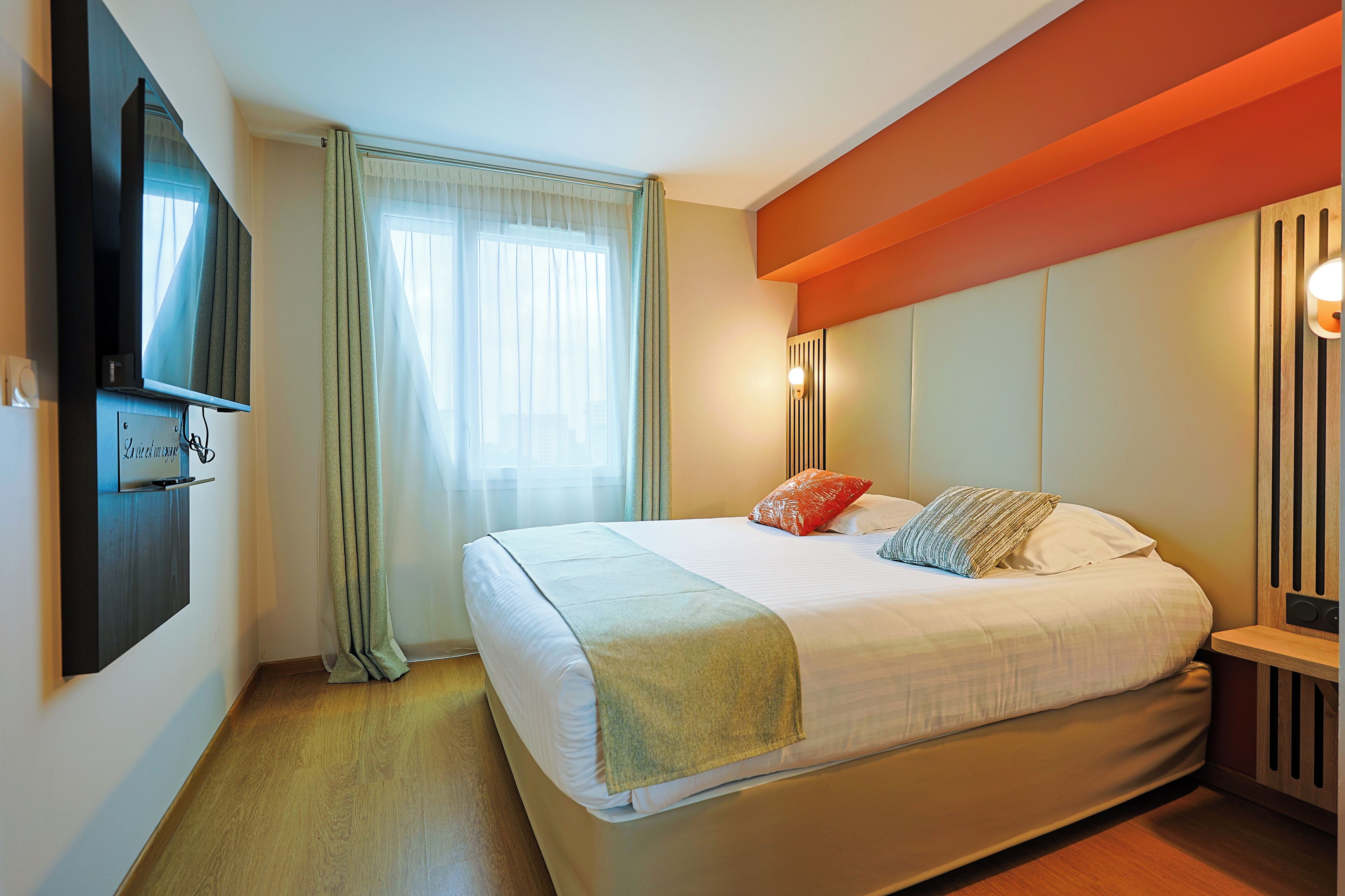 Un lit bien fait, avec une couette blanche et plusieurs oreillers, un téléviseur à écran plat fixé au mur, une fenêtre avec des rideaux légers et une table de chevet en bois avec une lampe.