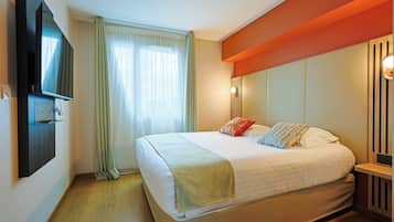 Un lit bien fait, avec une couette blanche et plusieurs oreillers, un téléviseur à écran plat fixé au mur, une fenêtre avec des rideaux légers et une table de chevet en bois avec une lampe.