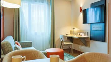 A modern hotel room with a sofa, a small table with two mugs, a desk with a chair, and a TV mounted on the wall.