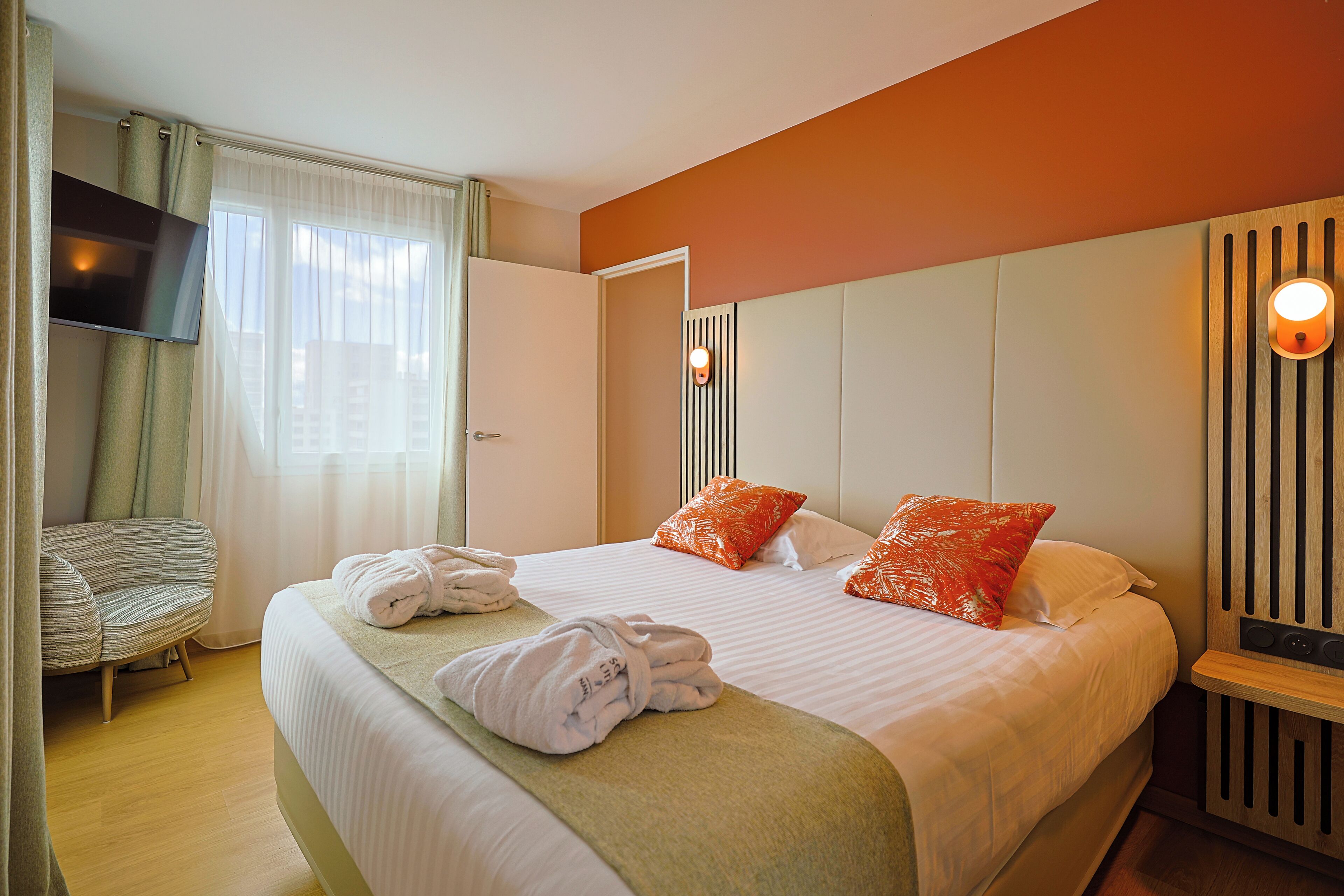 Una habitación de hotel con una cama, televisor, una silla y una mesita de noche con lámpara.