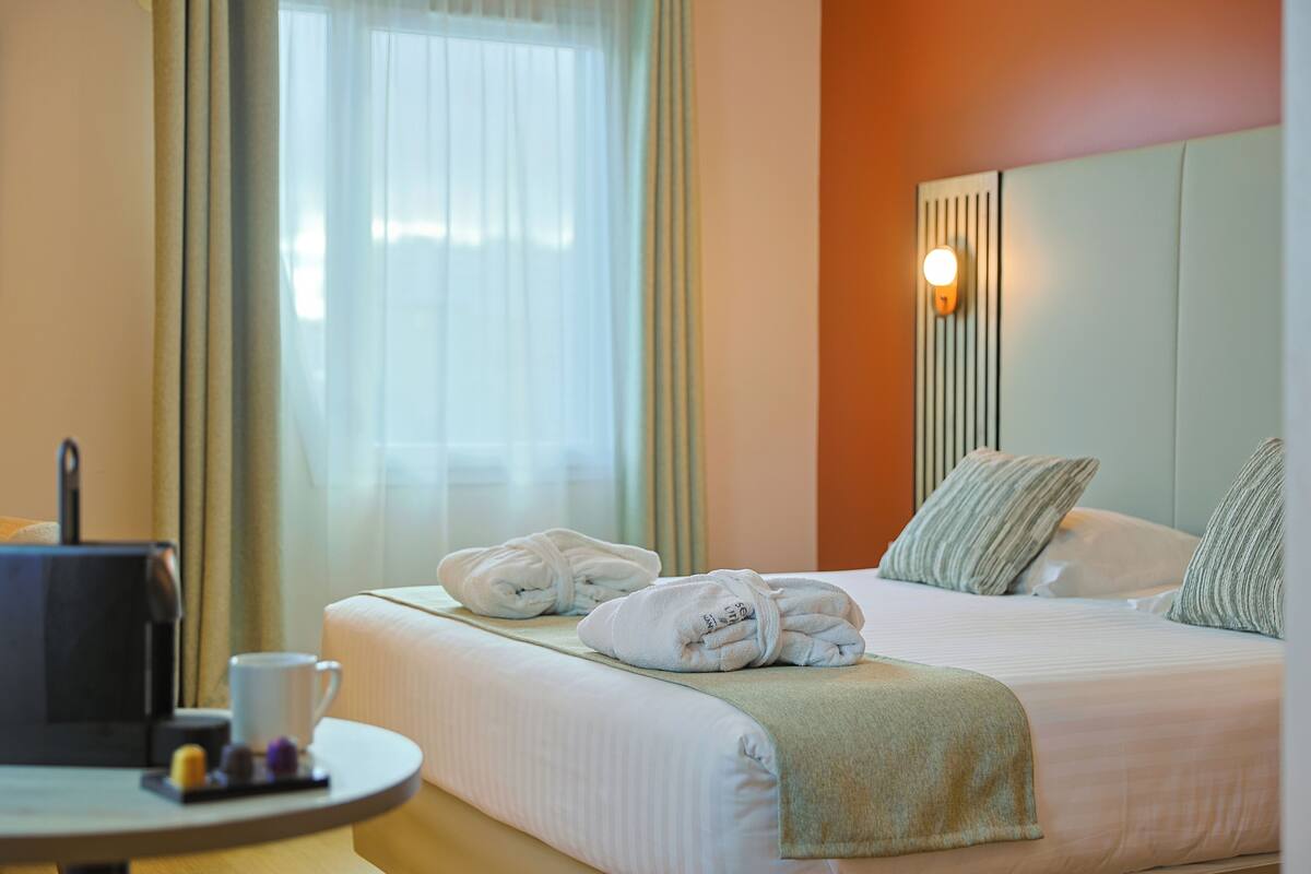 Een hotelkamer met een bed, kussens, een nachtkastje met een koffiezetapparaat en een raam met gordijnen.
