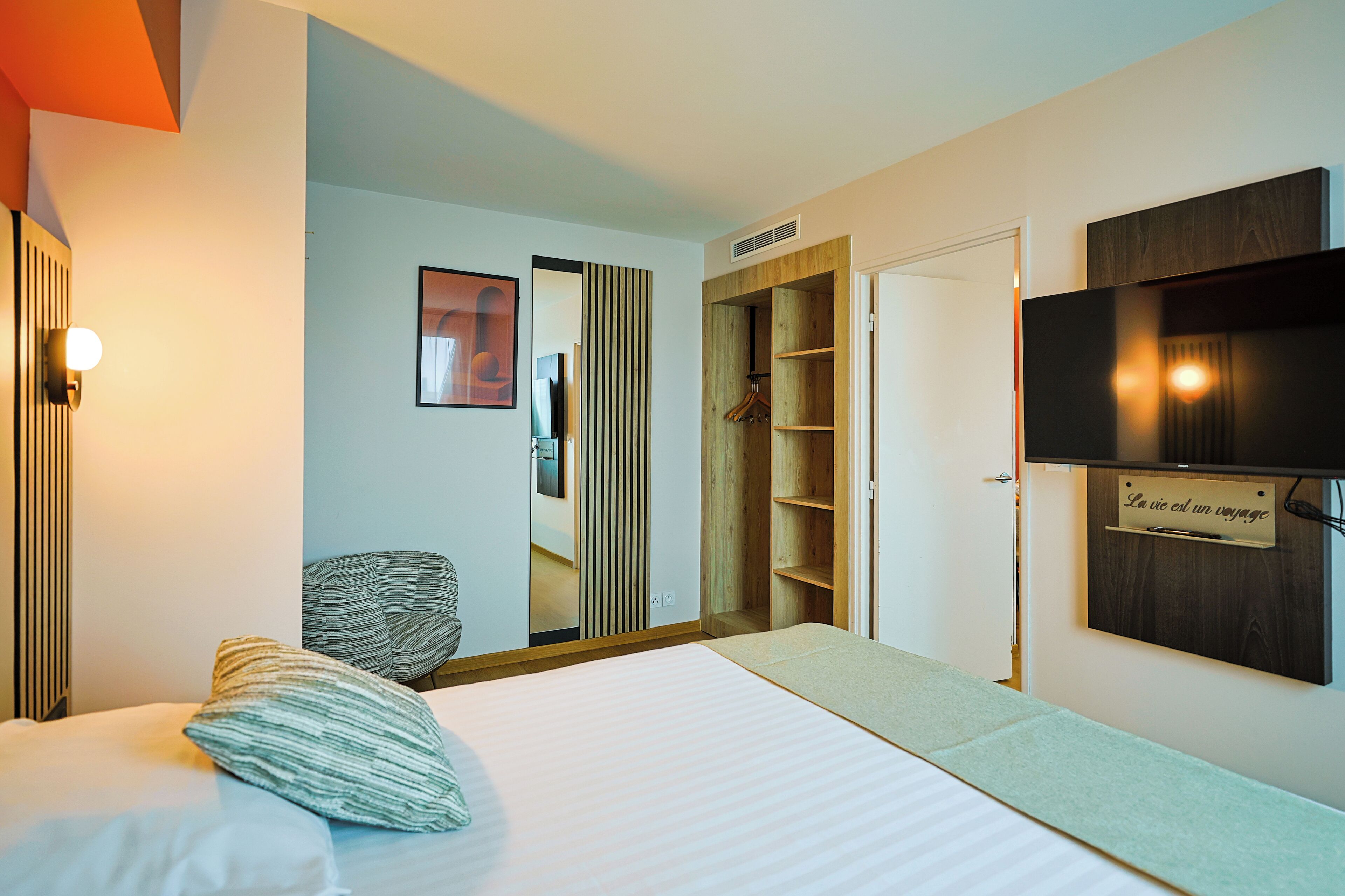 A modern hotel room with a bed, a chair, a wardrobe, and a mounted TV.