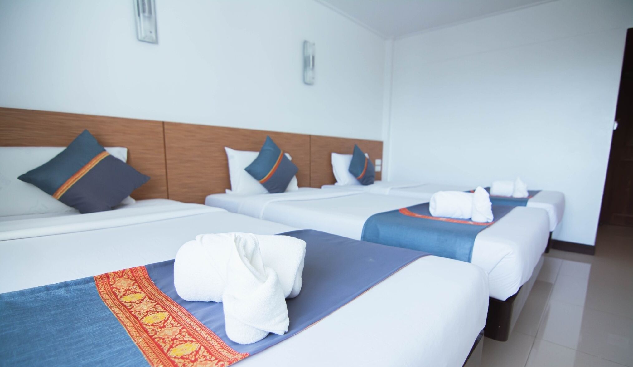 Ein Hotelzimmer mit drei Betten, jedes mit einer blauen Bettdecke und weißen Kissen. Auf den Betten liegen Handtücher und es gibt ein Holz-Kopfteil.
