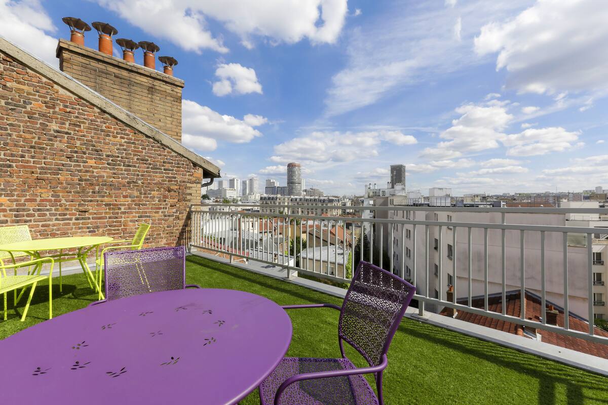 A rooftop terrace with a purple table and chairs, a brick wall, and a city view.