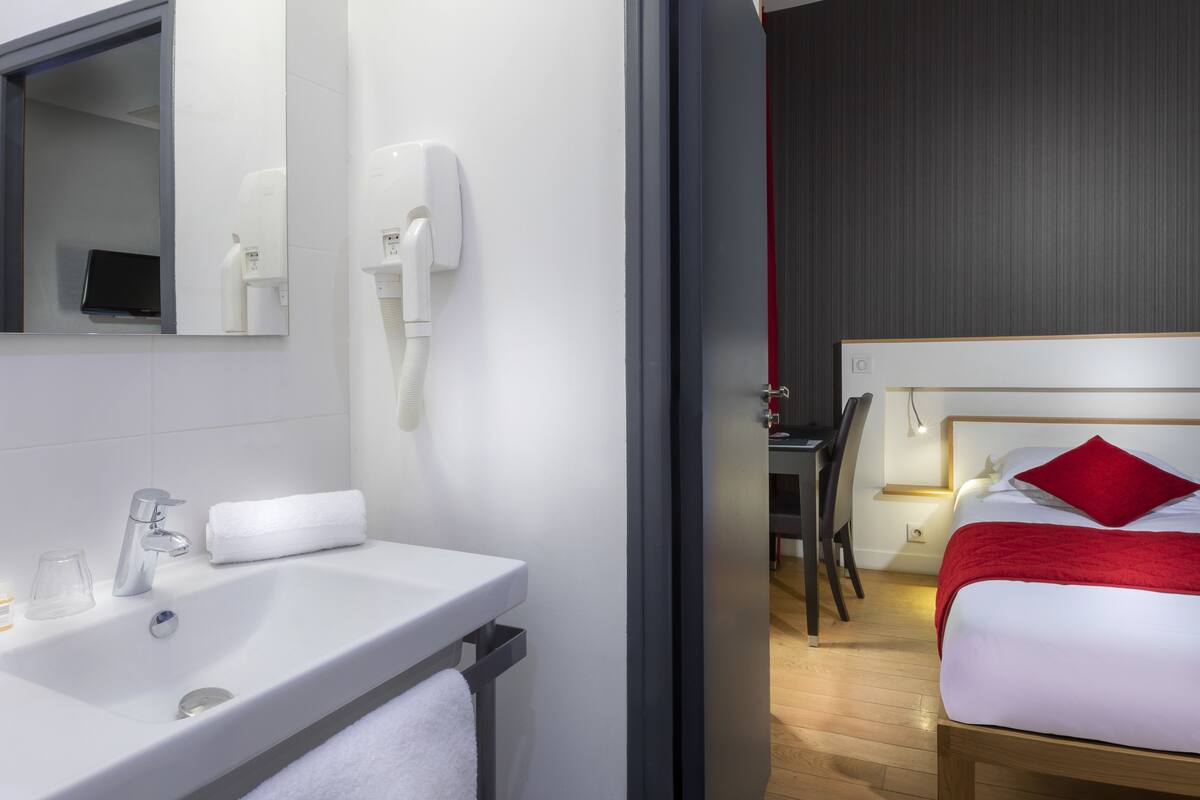 A modern hotel room with a white sink, a mirror, a hairdryer, a desk, and a bed with a red pillow.