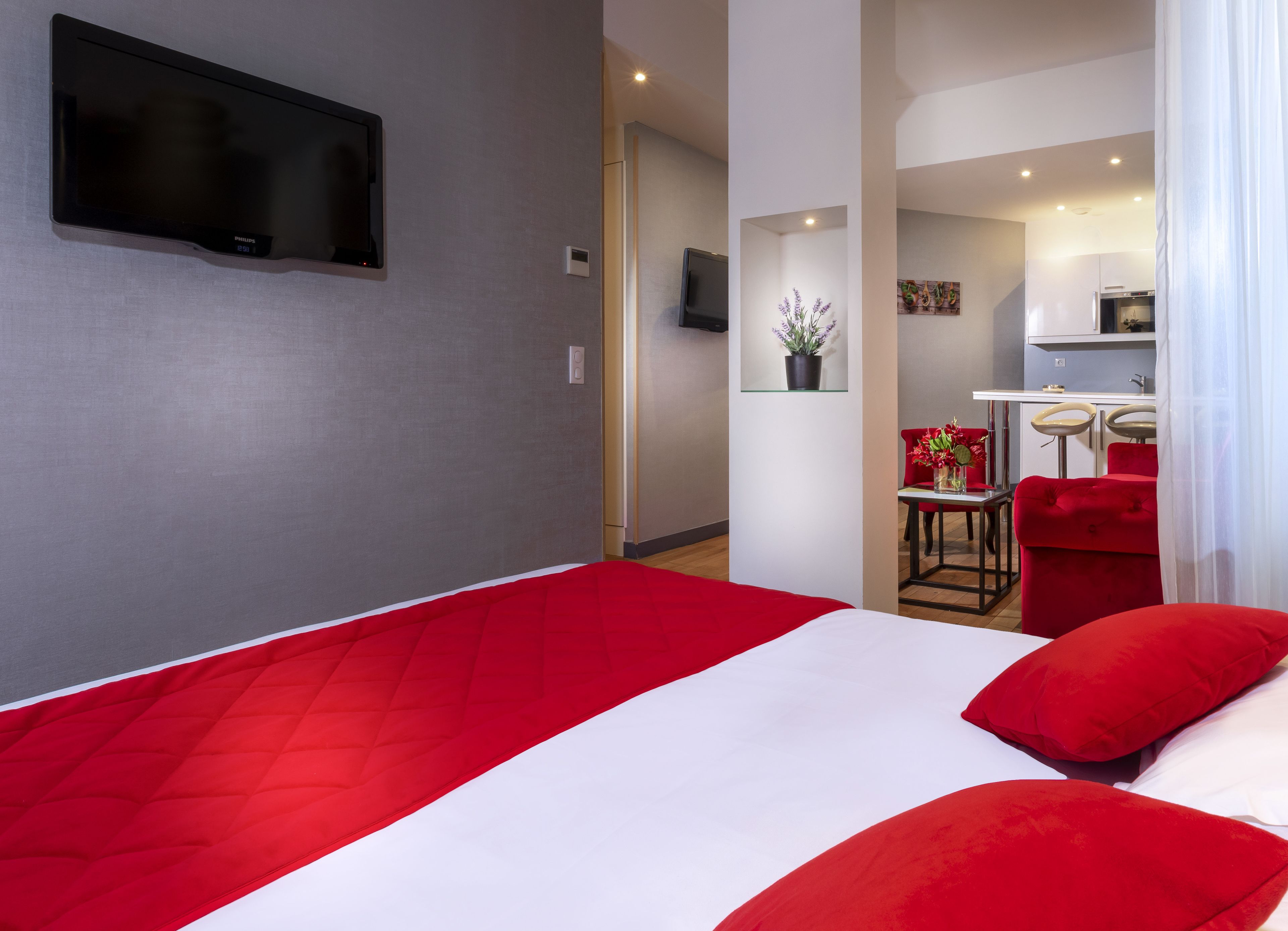 A hotel room with a large bed, a flat-screen TV mounted on the wall, a small kitchenette, and a red chair.