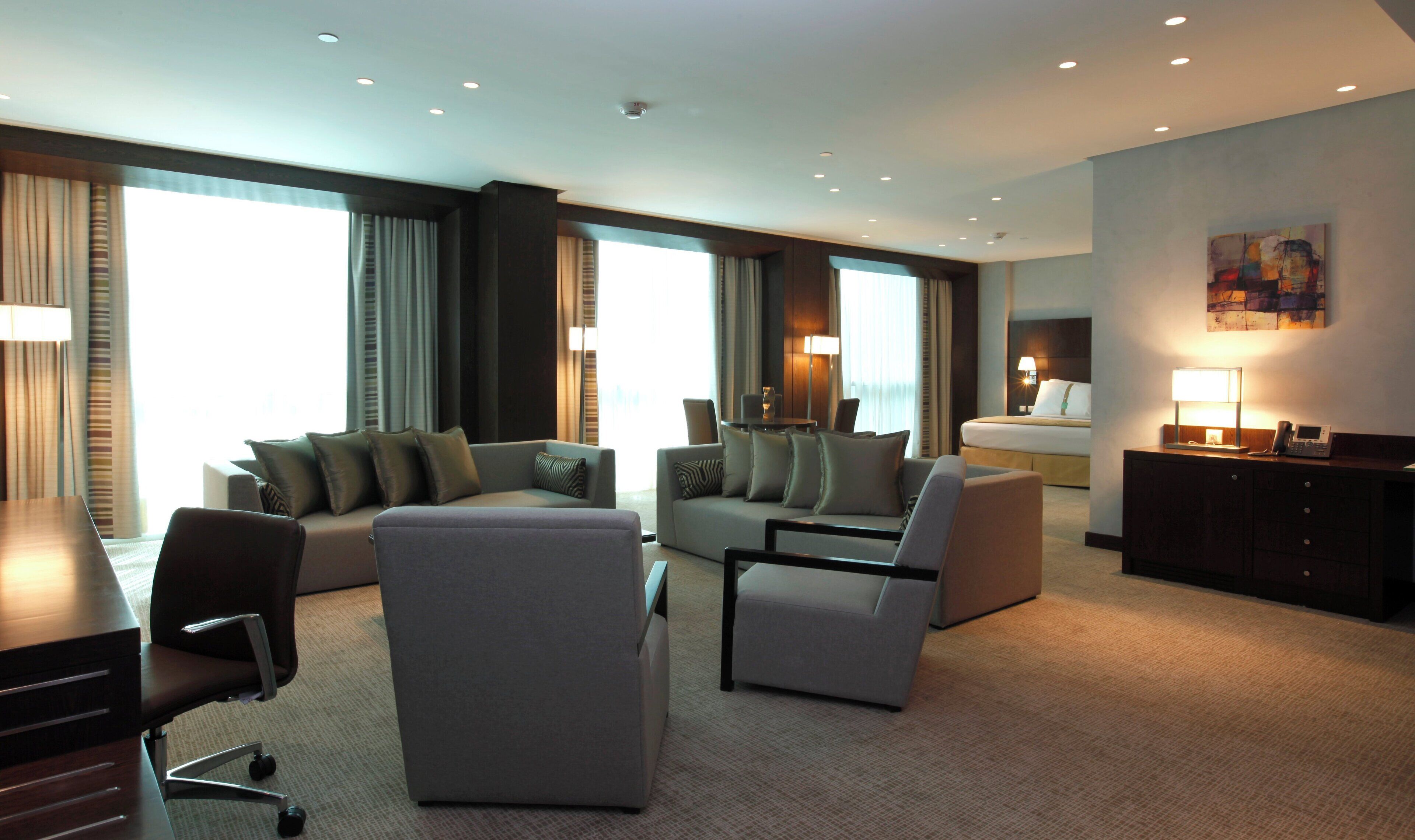 A modern hotel room with a living area featuring sofas, a coffee table, and a desk with a chair. There is a bed with a bedside table and lamp, and a painting on the wall.