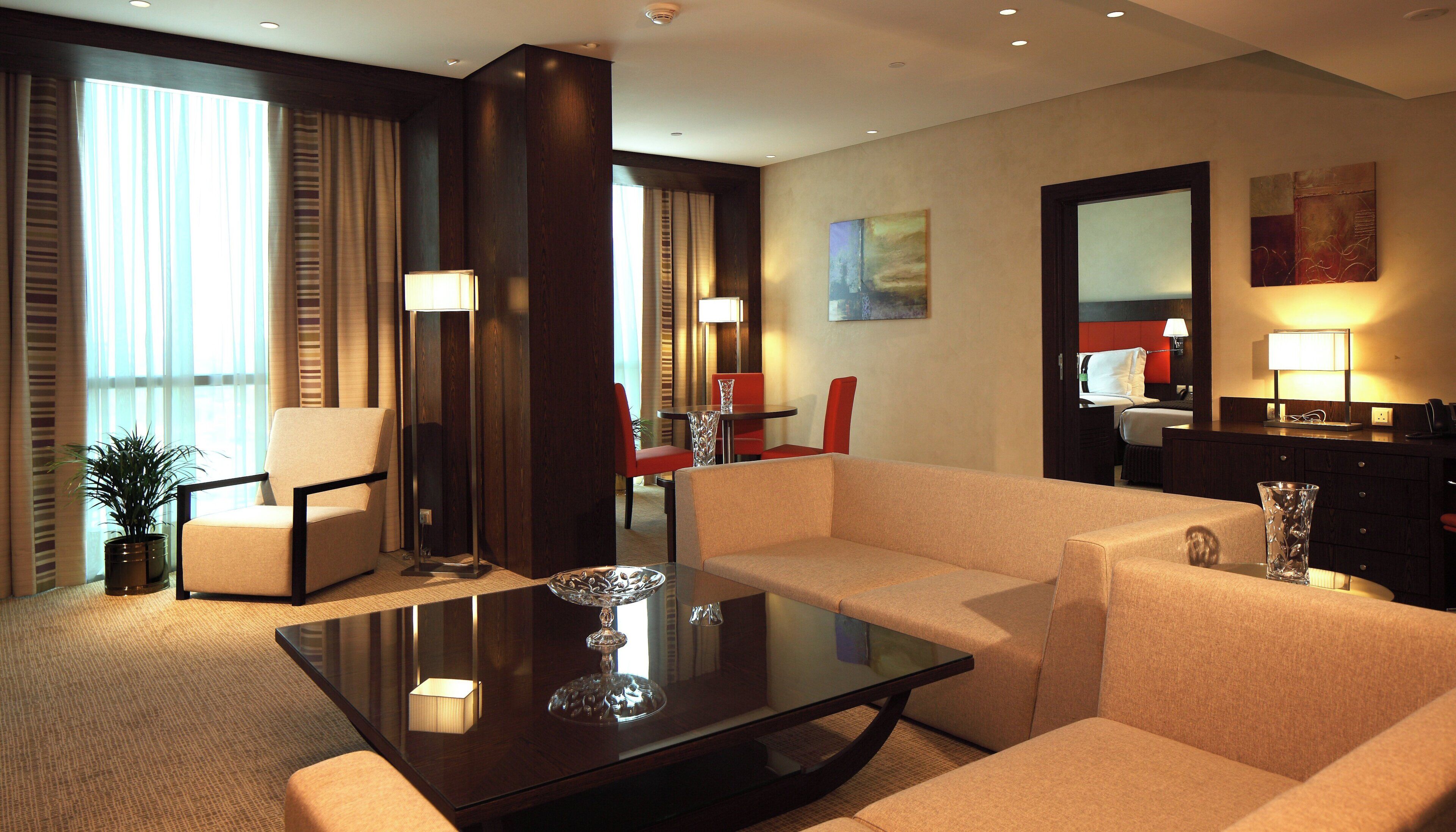 Una habitación de hotel con sofá, una mesa de centro de vidrio, una mesa de comedor con sillas, una lámpara y un ventanal con cortinas.