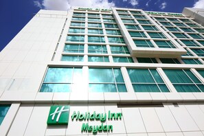 Een Holiday Inn Meydan hotelgebouw met een modern architectuurontwerp, met een opvallend groen logo en een helder uitzicht op de lucht.