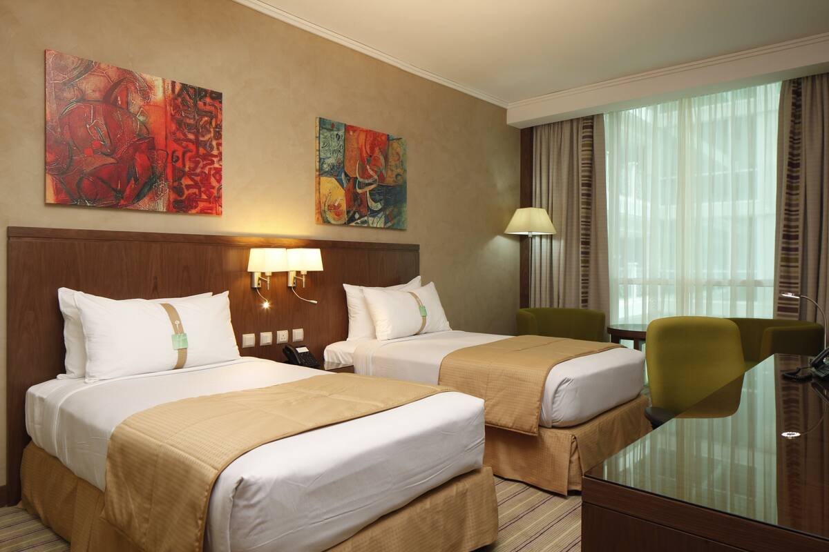 A hotel room with two beds, a desk, and a chair. There are two paintings on the wall, a lamp, and a window with curtains.