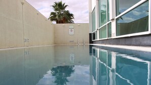 A clear indoor swimming pool with a no diving sign, a palm tree, and a building with large windows in the background.