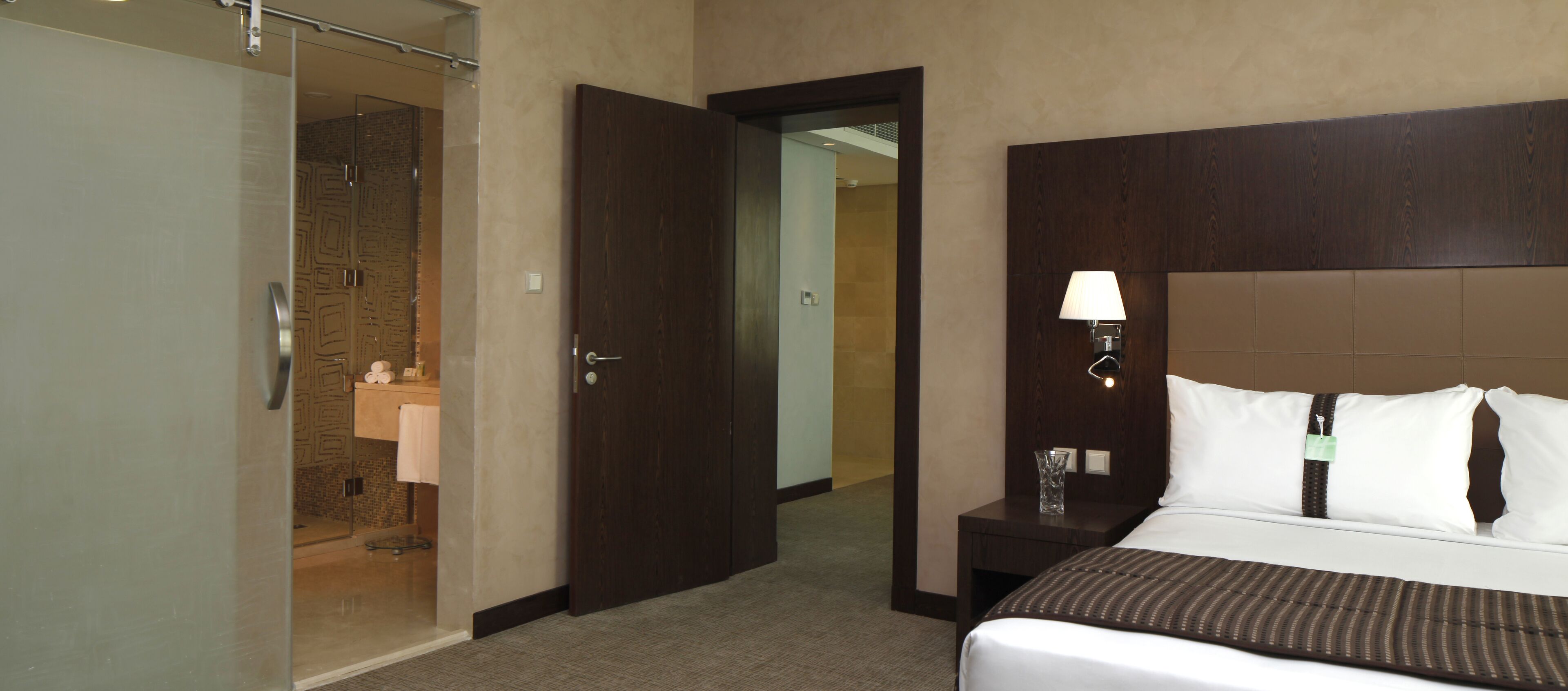 Ein Hotelzimmer mit Bett, Nachttisch, Lampe, Bad mit Dusche und einer Tür zu einem weiteren Raum.